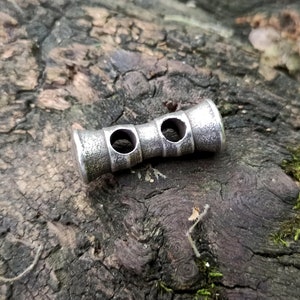 May include: A silver metal toggle with two holes, designed for use in clothing or accessories.