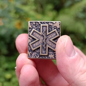 Pode incluir: Um alfinete de metal de cor bronze com uma superf&iacute;cie texturizada. O alfinete apresenta uma estrela de seis pontas com um s&iacute;mbolo m&eacute;dico no centro. A estrela &eacute; cercada por uma moldura quadrada.