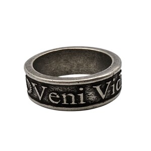 May include: A silver-toned metal ring with a weathered, dark finish. The band features the embossed Latin text Veni Vidi Vici in a serif font, with a small laurel wreath icon visible at the start of the inscription on the outer surface of the band.