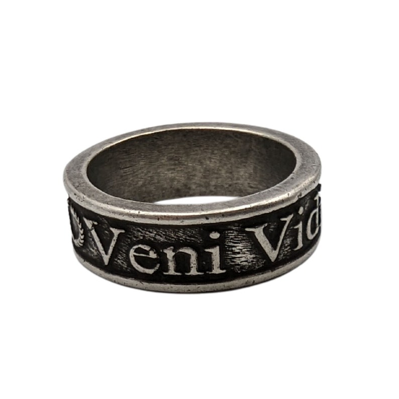 May include: A silver-toned metal ring with a weathered, dark finish. The band features the embossed Latin text Veni Vidi Vici in a serif font, with a small laurel wreath icon visible at the start of the inscription on the outer surface of the band.