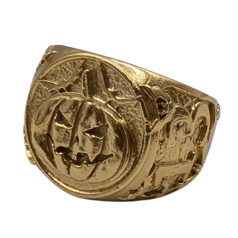 Puede incluir: Un anillo de color dorado con una talla detallada de una cara de calabaza de Halloween. El anillo es grueso y tiene una banda ancha.