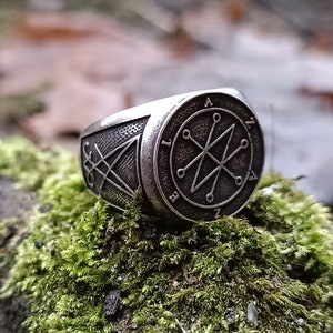 A silver ring with a black sigil design. The sigil is surrounded by a circle with Azazel name inside