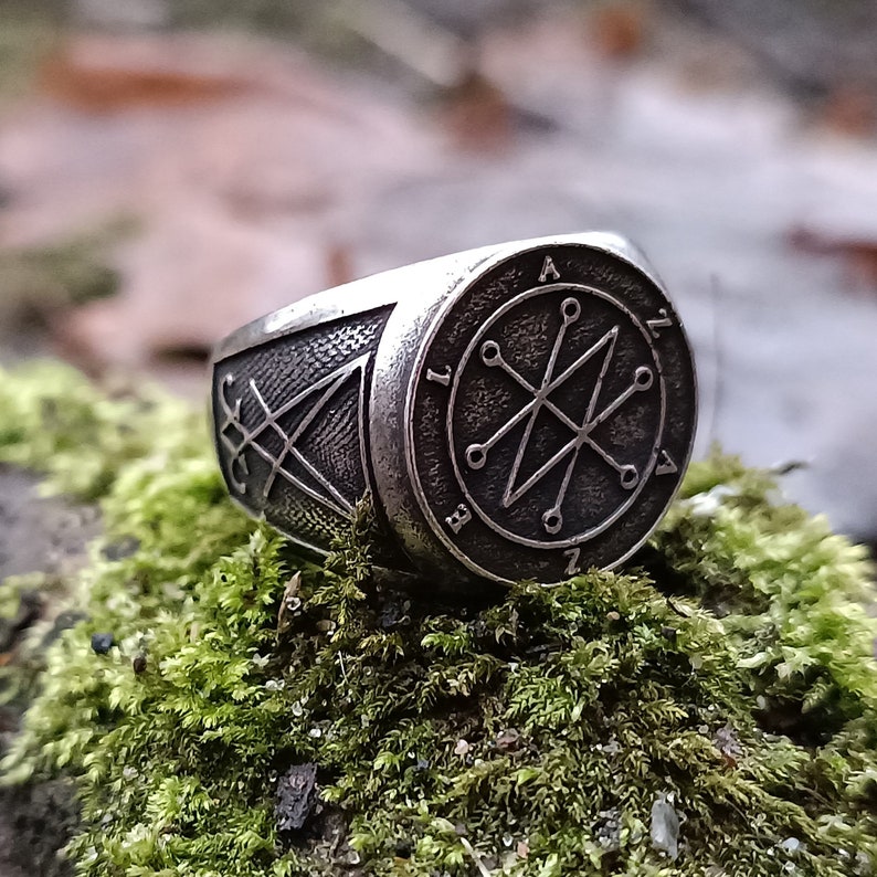 A silver ring with a black sigil design. The sigil is surrounded by a circle with Azazel name inside