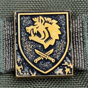 Puede incluir: Pin rectangular con borde dorado y fondo negro. El pin presenta un emblema dorado de la cabeza de una bestia gruñona sobre dos cuchillos cruzados. El pin está colocado sobre una tela verde y gris.