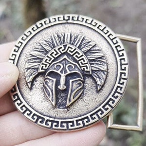 May include: A circular brass-toned metal belt buckle featuring a raised relief of a Spartan-style helmet centred within a border of Greek key patterns. The textured finish highlights the intricate details of the helmet crest and visor design.