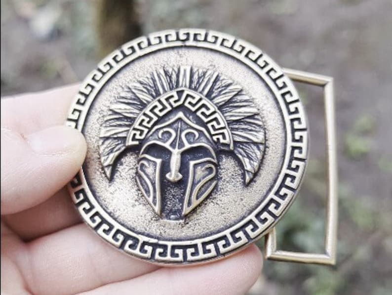 May include: A circular brass-toned metal belt buckle featuring a raised relief of a Spartan-style helmet centred within a border of Greek key patterns. The textured finish highlights the intricate details of the helmet crest and visor design.