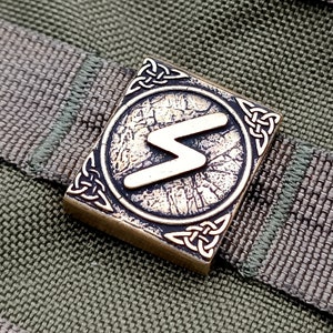 May include: A square bronze-toned metal buckle featuring a central circular emblem with a raised lightning bolt symbol. The corners of the buckle are decorated with intricate Celtic knotwork patterns, and it is attached to a textured olive green strap.