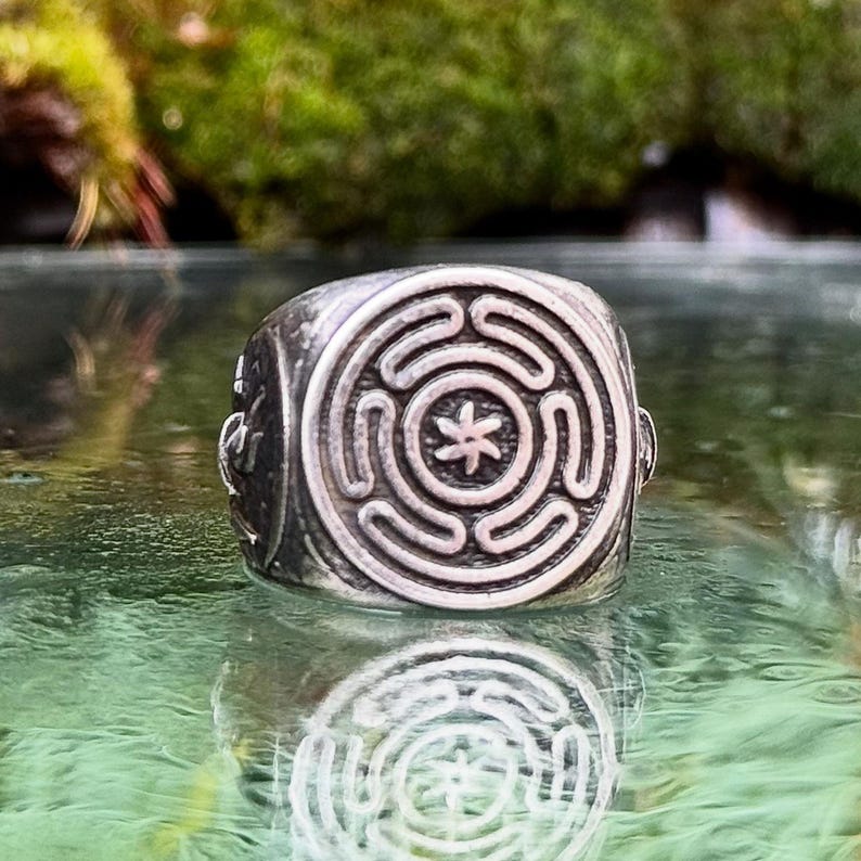 May include: A silver-toned metal ring featuring a circular face with an engraved labyrinth pattern and a central six-pointed star symbol. The band has a weathered, dark finish with additional etched details visible on the side of the ring structure.