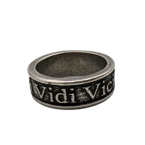 May include: A silver-toned metal ring with a weathered, dark finish. The band features the embossed Latin text Veni Vidi Vici in a serif font. The surface of the ring has a textured, distressed appearance with raised lettering and recessed dark areas.