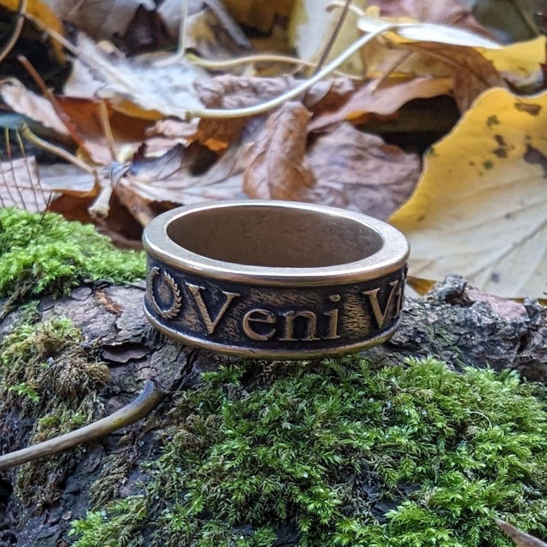 May include: A metallic band ring with a dark, weathered finish featuring the embossed Latin text Veni Vidi Vici and a laurel wreath symbol. The ring has a wide, flat profile with raised lettering and a textured, antique-style appearance.