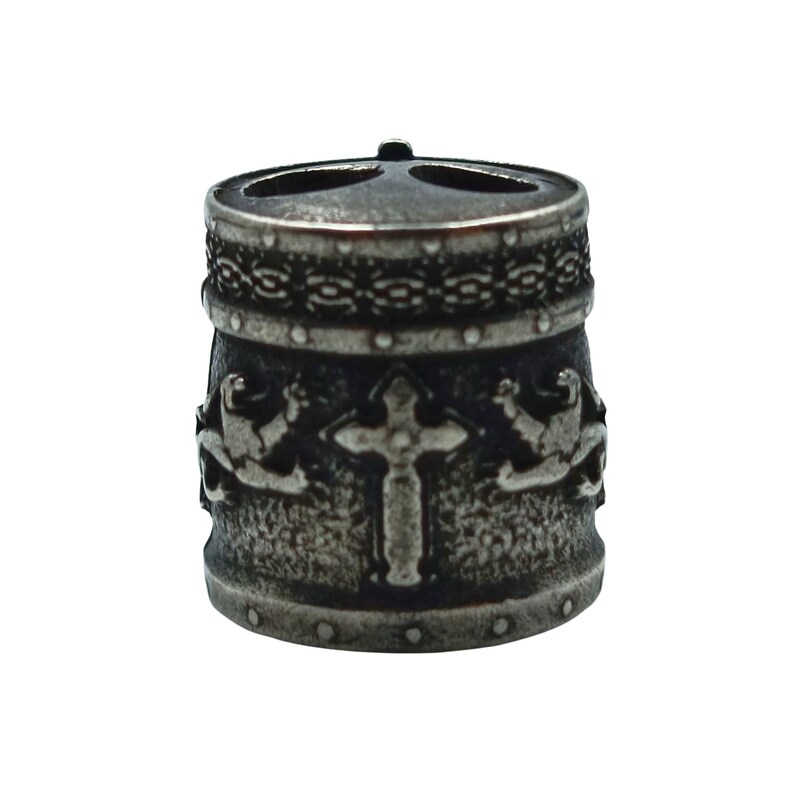 May include: A silver metal bead with a cross and two winged figures on the front. The bead has a textured surface and a decorative band around the top.