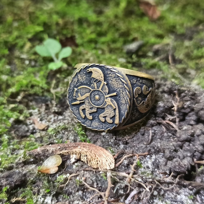 May include: A gold-toned ring with a detailed design of a warrior on horseback. The ring is set against a background of green moss.