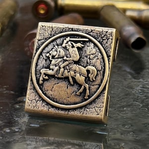May include: A rectangular, bronze-colored metal bead with a detailed relief of a knight on horseback within a circular frame. The bead has a textured, aged appearance, suggesting a vintage or antique style.