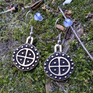 May include: A pair of silver earrings with a bronze circular pendant featuring a cross with four dots in each quadrant. The pendant is surrounded by a circle of small dots.