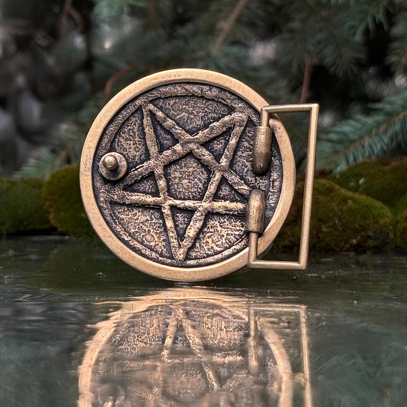 May include: A circular bronze-toned belt buckle featuring a raised pentagram design with a textured, weathered finish. The buckle includes a rectangular loop and a small metal stud on the side for attachment, showcasing a metallic antique appearance.