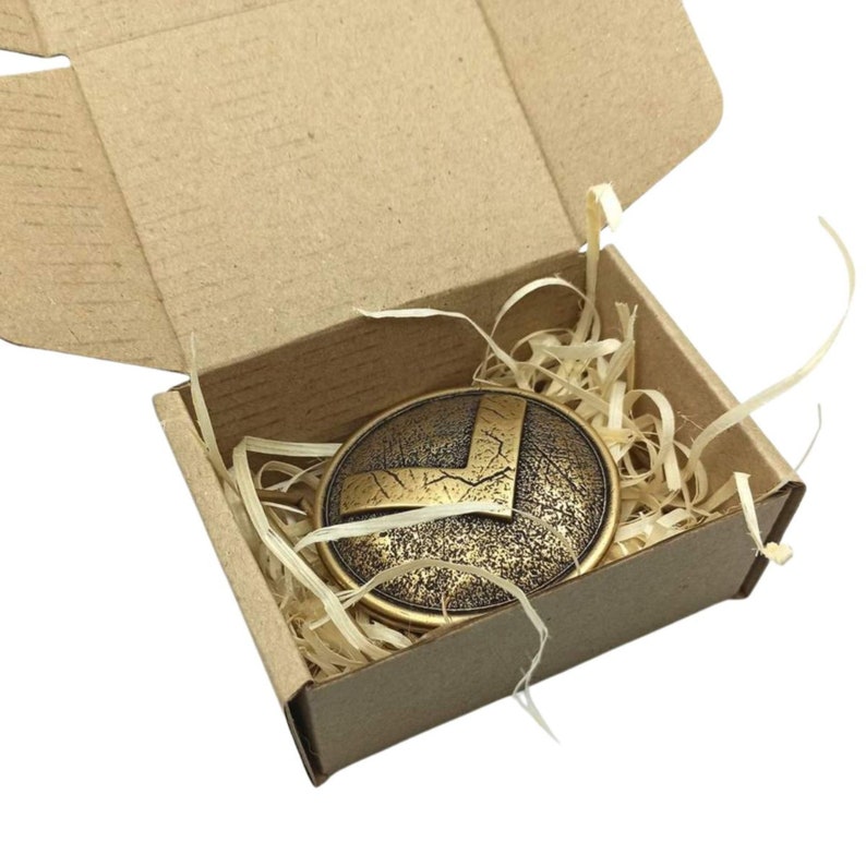 May include: A circular bronze-toned metal emblem featuring a raised V-shaped design with a distressed, textured finish. The emblem is nestled inside a small, open rectangular cardboard box filled with light-coloured wood shavings for padding.