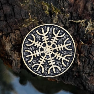 May include: A circular pendant with a bronze finish, displaying the Vegvísir symbol, a Viking compass. The pendant has a black background with gold-coloured lines. The background is a dark, textured surface.