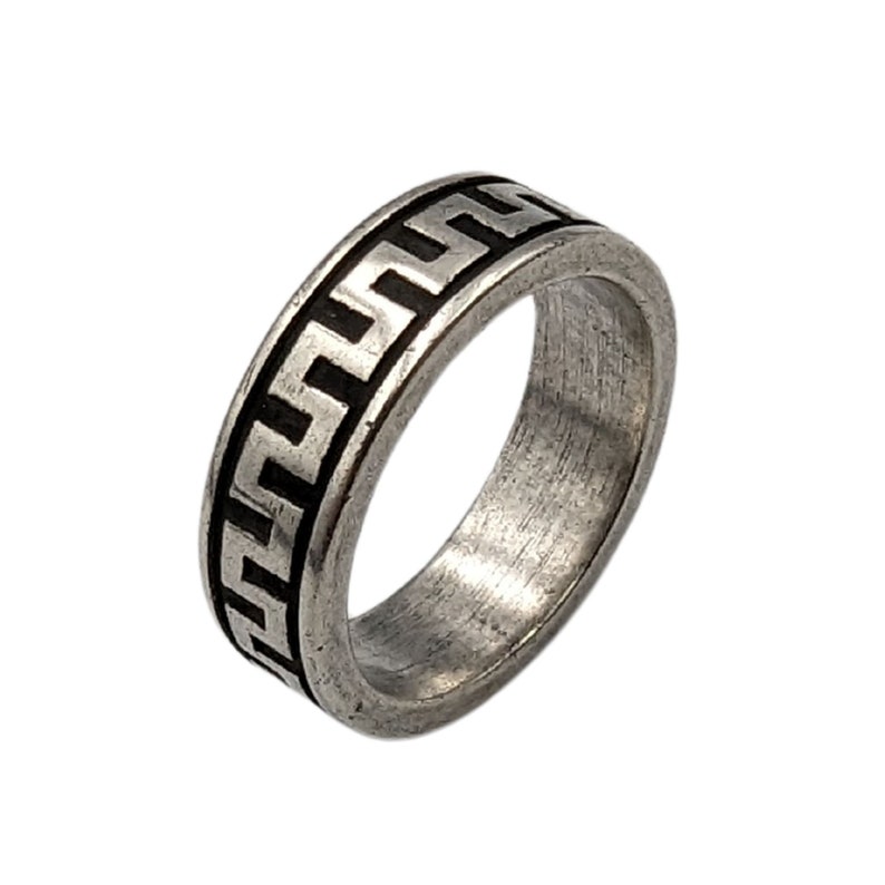 May include: A silver-toned metal ring featuring a continuous black recessed Greek key pattern around the outer band. The surface of the ring has a brushed, matte finish with visible texture, creating a contrast between the dark geometric design and the metallic band.