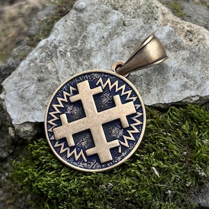 May include: A bronze-colored pendant with a Jerusalem cross design. The cross is centered within a circular frame with a zigzag border. The pendant has a loop for a chain and rests on a mossy surface.