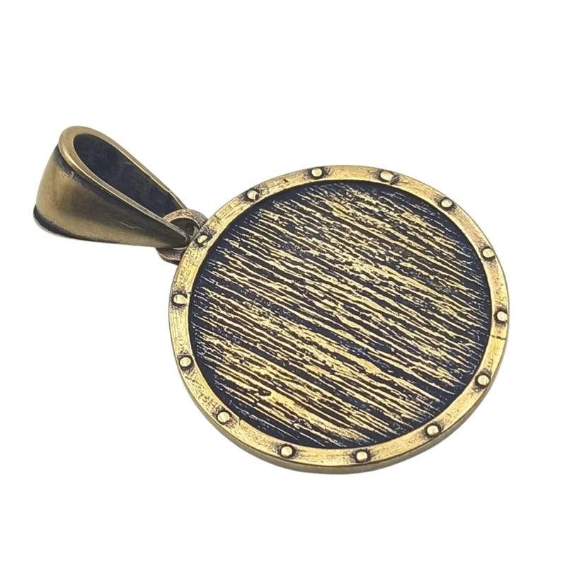May include: A round, gold-toned pendant with a textured surface and a decorative border. The pendant has a loop at the top for attaching to a chain.