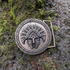 May include: A circular bronze-toned belt buckle featuring a raised relief of a Spartan-style helmet with a plume. The outer edge of the buckle is decorated with a continuous Greek key pattern, and a rectangular loop is attached to the right side.
