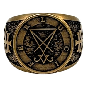 May include: A bronze-toned metal ring featuring a circular face with a textured dark background. The center displays a sigil of Lucifer, surrounded by the letters L, U, C, I, F, E, R. The sides of the ring band are adorned with embossed cross symbols.