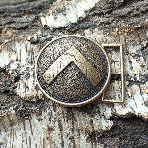 May include: A round, bronze-toned metal belt buckle featuring a raised, textured inverted V-shaped emblem in the centre. The surface of the buckle has a distressed, weathered finish with visible scratches and dark oxidation in the recessed areas.