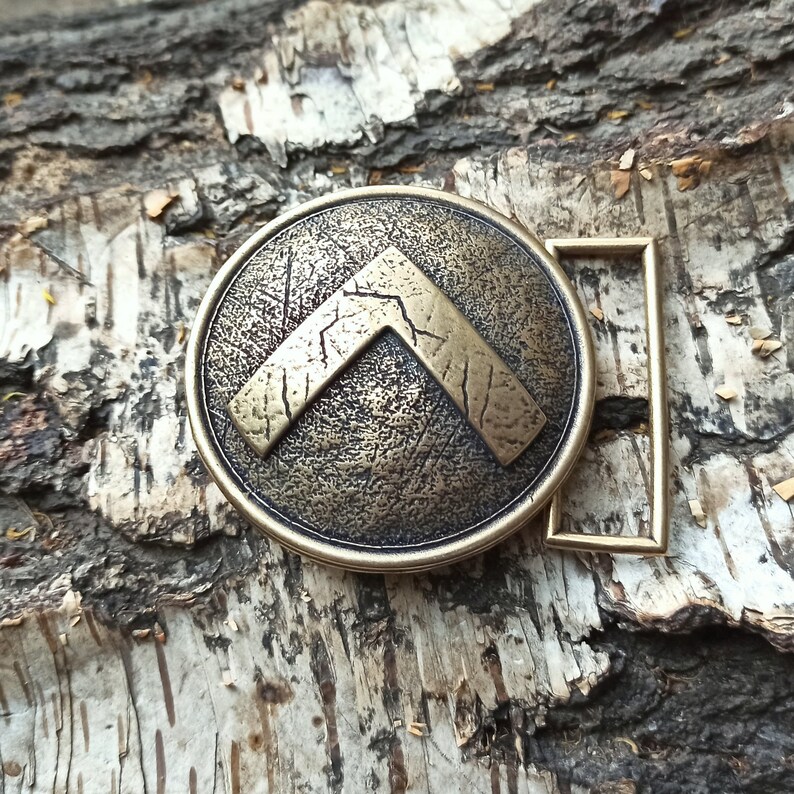 May include: A round, bronze-toned metal belt buckle featuring a raised, textured inverted V-shaped emblem in the centre. The surface of the buckle has a distressed, weathered finish with visible scratches and dark oxidation in the recessed areas.