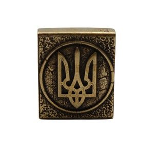 May include: A gold-colored metal square with a textured surface. The square features a raised, circular design with a stylized trident, the national symbol of Ukraine, in the center.