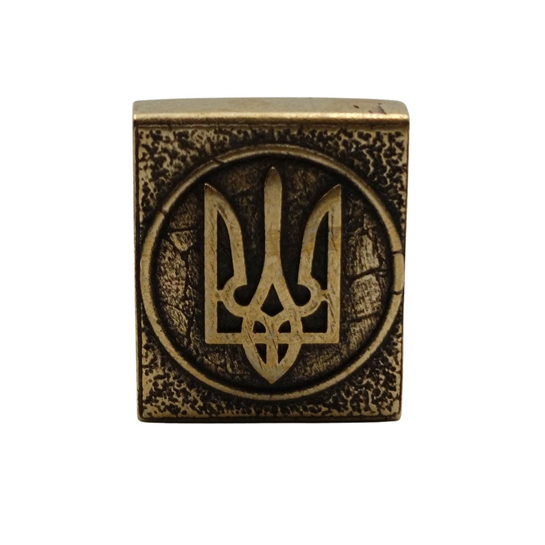 May include: A gold-colored metal square with a textured surface. The square features a raised, circular design with a stylized trident, the national symbol of Ukraine, in the center.