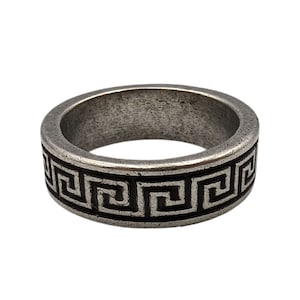 May include: A silver-toned metal ring featuring a continuous black recessed Greek key pattern around the outer band. The surface of the metal has a weathered, matte finish with visible texture and a rounded profile for the band.