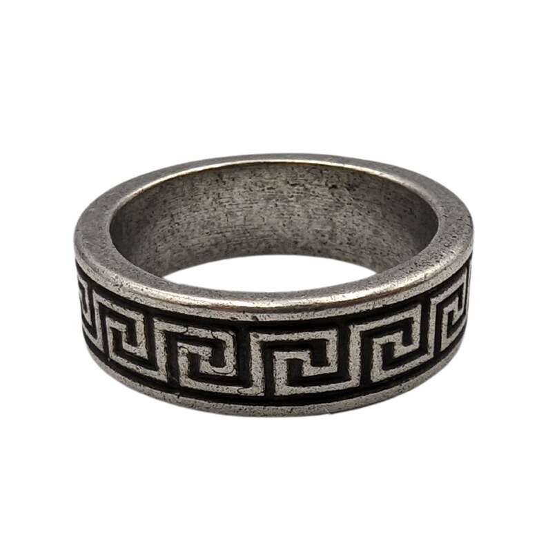 May include: A silver-toned metal ring featuring a continuous black recessed Greek key pattern around the outer band. The surface of the metal has a weathered, matte finish with visible texture and a rounded profile for the band.