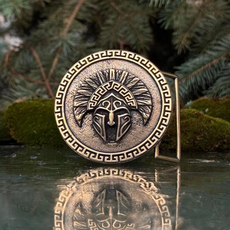 May include: A circular bronze-toned belt buckle featuring a central embossed Spartan helmet design. The outer border is decorated with a repeating Greek key pattern. The metal surface has a textured, weathered finish with dark recessed details.