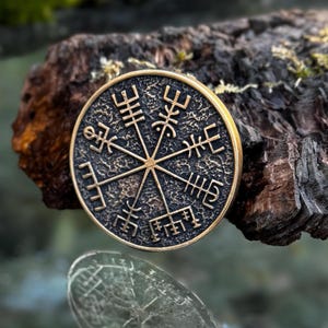 Pode incluir: Um pingente redondo, cor de bronze, com um design detalhado de bússola viking. O pingente tem um fundo escuro e texturizado e uma borda dourada. O design da bússola é uma série de linhas e símbolos que se cruzam.