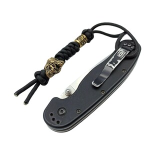 May include: A black folding knife with a black paracord lanyard featuring a skull bead and a metal bead with a geometric design.
