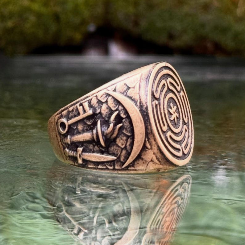 May include: A bronze-toned metal ring featuring an engraved circular labyrinth design on the face. The side of the band displays embossed imagery of a crescent moon, a key, a torch, and a dagger set against a textured, stone-like background.