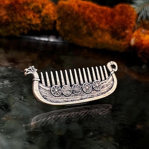 Bronze beard comb Viking ship Drakkar comb with intricate detailing. The comb features a dragon head at the front and a curved design at the back. Shields with various symbols are etched along the ship's side. The comb's teeth are evenly spaced.