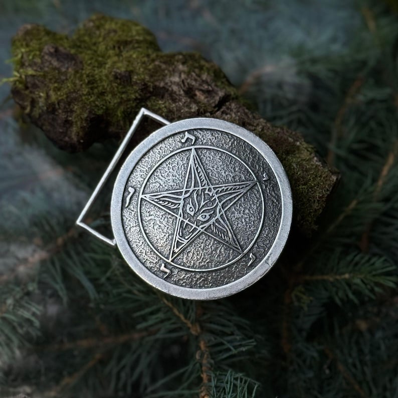 May include: A circular silver-toned metal belt buckle featuring a textured surface with an embossed five-pointed star design. Inside the star is a stylized animal head, and the outer border contains small, etched symbols resembling ancient runes.