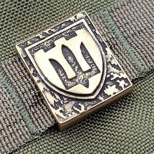 Könnte beinhalten: Ein goldfarbener Metallaufnäher mit einem stilisierten Dreizack-Design. Der Aufnäher hat einen strukturierten, Tarnmuster-ähnlichen Hintergrund.