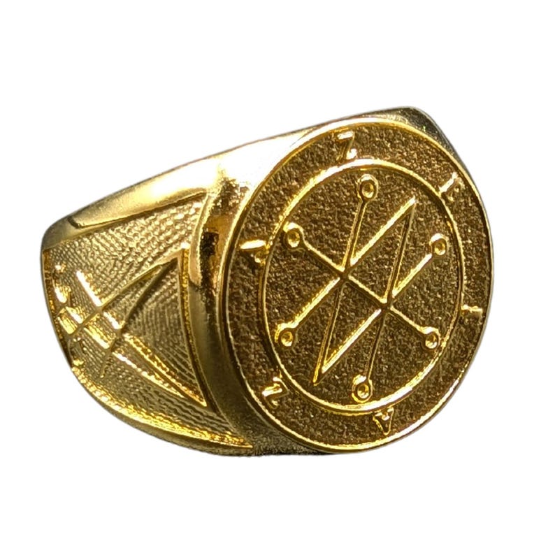 A gold Azazel sigil ring with a circular design featuring a starburst pattern and a circle around the edge. The ring is made of metal and has a smooth, polished finish.
