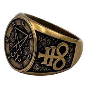 May include: A brass-toned metal ring featuring a circular face with the engraved sigil of Lucifer and the letters F, E, R, L, U, C, I arranged around the border. The side of the ring displays a raised, textured black background with a gold symbol.