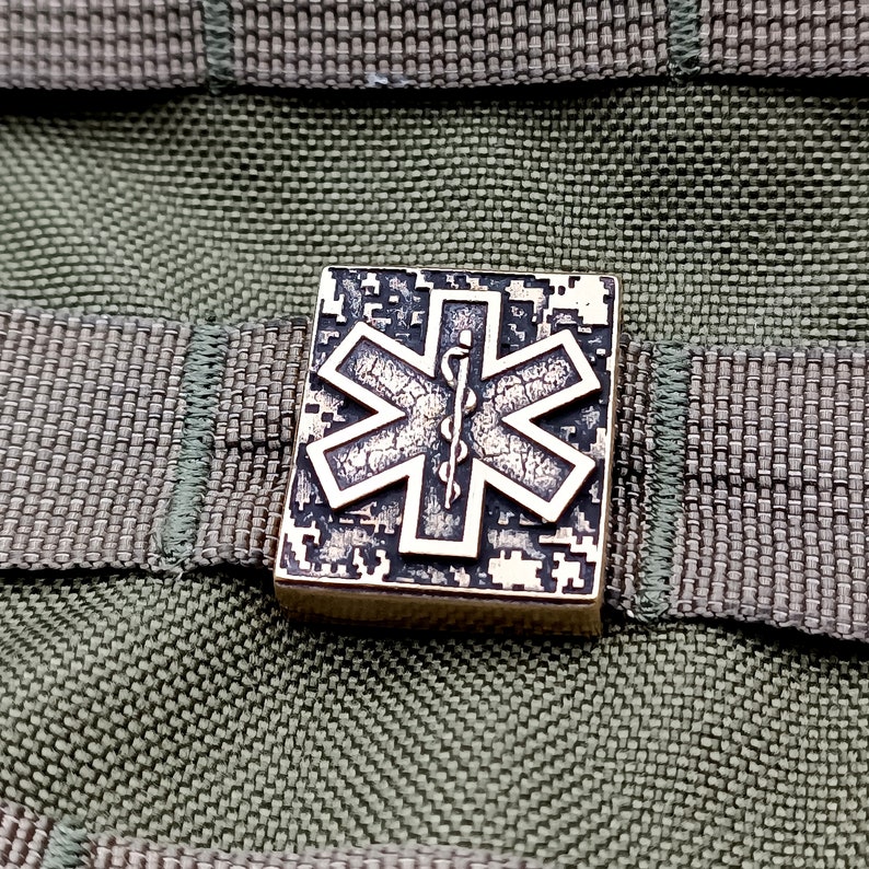 Pode incluir: Um remendo quadrado de camuflagem com um s&iacute;mbolo m&eacute;dico prateado, a Estrela da Vida. O remendo &eacute; de metal e tem uma superf&iacute;cie texturizada.