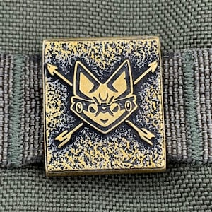May include: A rectangular, gold-colored metal pin featuring a stylized cat face with crossed arrows. The pin has a textured background and is set against a green fabric background. The cat face has large eyes and pointed ears.