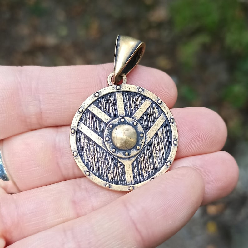 May include: A gold-toned pendant in the shape of a round shield with a textured surface. The shield has a central circular design with a raised, textured surface. The pendant is on a chain.