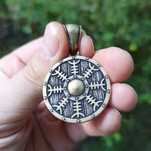 May include: A bronze pendant in the shape of a shield with a circular design. The shield has a textured surface and a raised centre with a circular design. The pendant is on a chain.