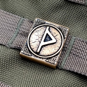 Pode incluir: Um amuleto de metal de cor bronze com um design de nó celta e um símbolo de runa dentro de um círculo. O amuleto está sobre um fundo de tecido verde.