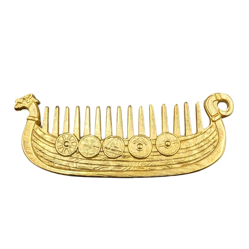 A gold plated beard comb shaped like a Viking ship Drakkar. The comb features a detailed design of a dragon head at the front and a circular loop at the back. The ship has a series of circular shields along the side and vertical teeth for combing hair or beard.