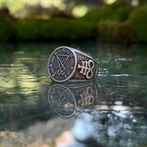 May include: A silver-toned metal ring featuring a circular face with an engraved sigil of Lucifer and the letters L U C I F E R around the perimeter. The side of the band displays a raised Leviathan cross symbol against a textured, dark background.