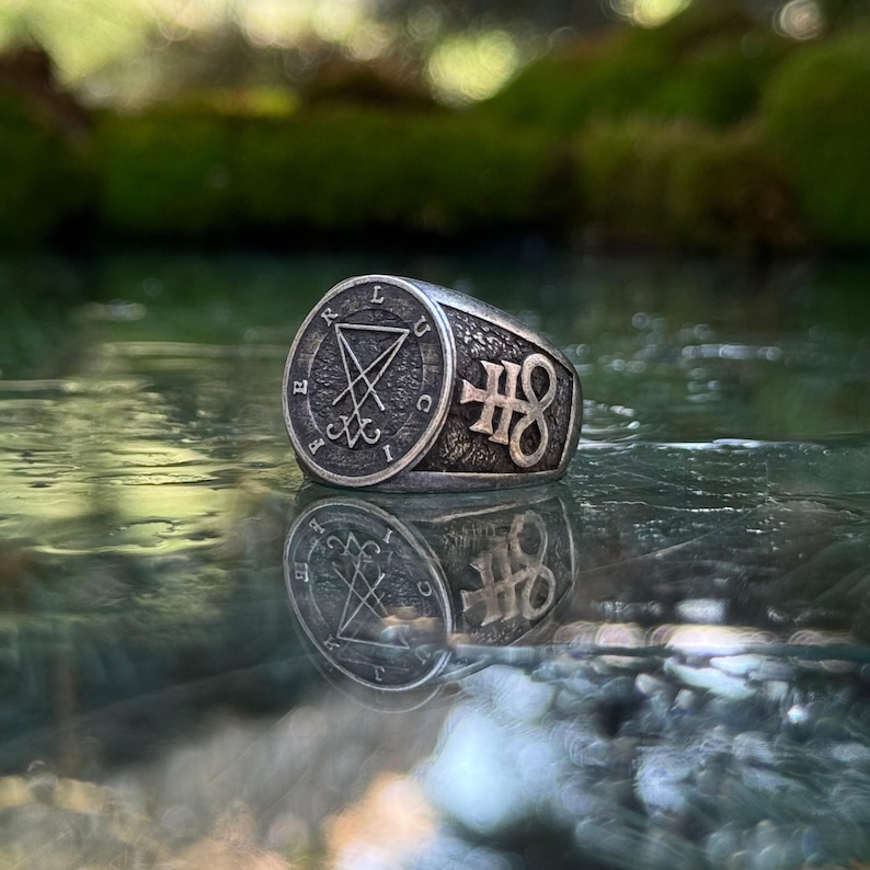 May include: A silver-toned metal ring featuring a circular face with an engraved sigil of Lucifer and the letters L U C I F E R around the perimeter. The side of the band displays a raised Leviathan cross symbol against a textured, dark background.