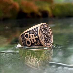 May include: A bronze-toned metal ring featuring a textured surface with a prominent Leviathan cross symbol on the side and a circular seal on the face containing a sigil and the letters L, U, C, I, F, E, R arranged around the perimeter.